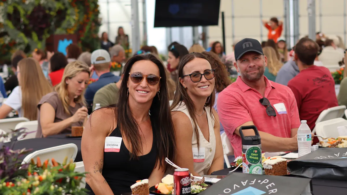 Three guests smiling at a lunch table in the Shipping Greenhouse, with the flower wall behind
